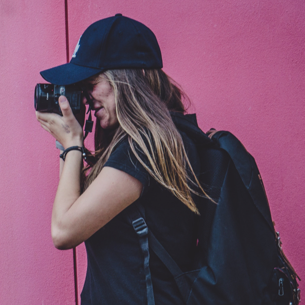girl wearing all black with a black hat and backpack - taking a photo in front of a pink wall