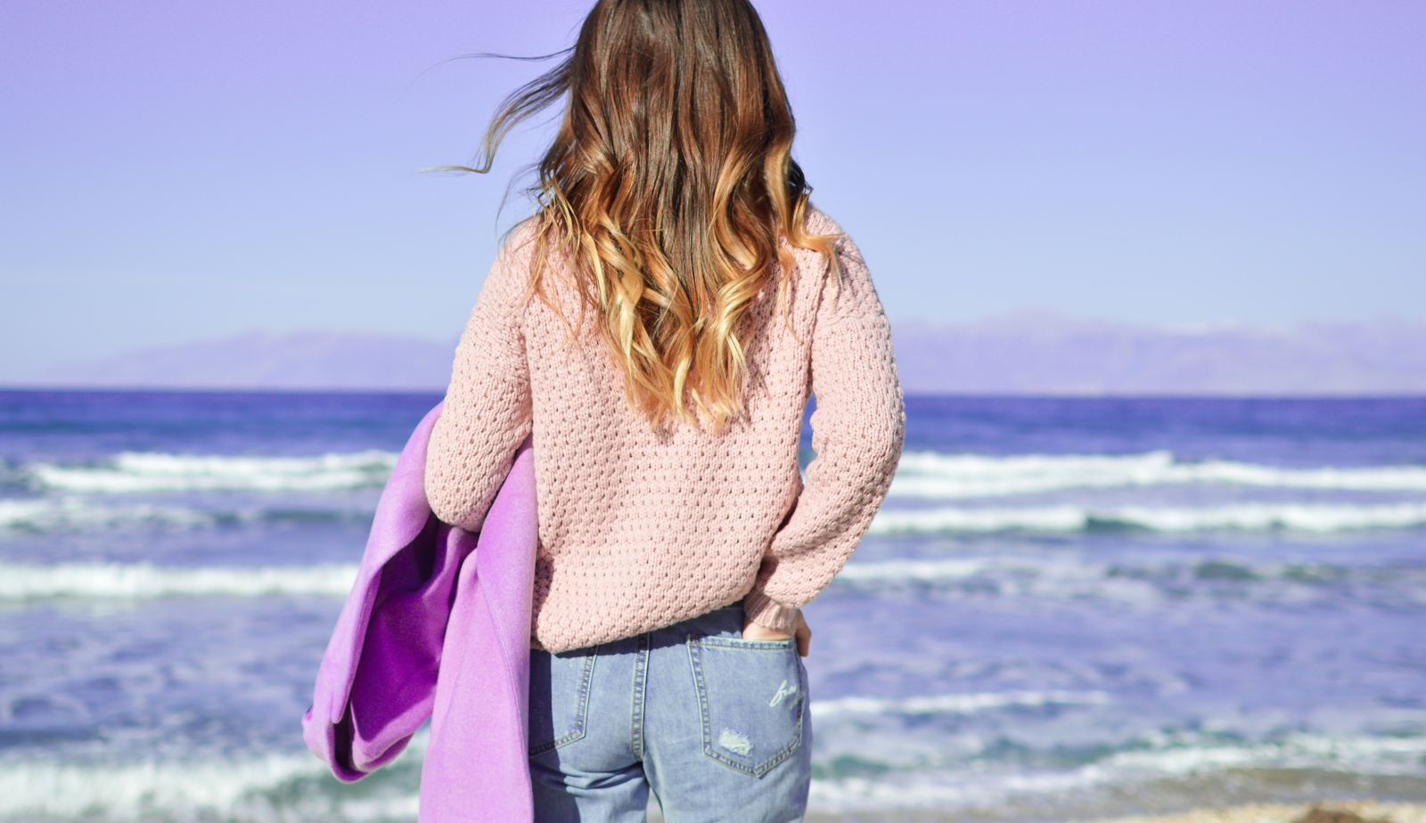 girl with hombre hair and a knit sweater walking towards the ocean holding a jean jacket